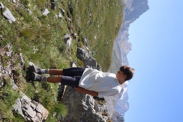 A young person stands on a rocky, grassy mountain slope, looking out towards distant peaks under a clear blue sky
